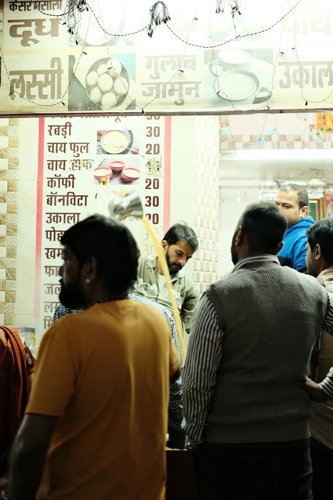 Customers at a busy tea and snacks counter with a Hindi menu board